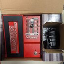 Červený efekt pedál s černým ovládacím pedálem v kartonové krabici. Na pedálu je nápis Whammy a ovládací panel s popisky HARMONY, WHAMMY, CLASSIC, CH…