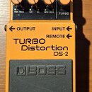 Oranžový kytarový efekt s černým pedalem. Na šasi jsou nápisy TURBO Distortion DS-2, OUTPUT, INPUT a REMOTE, v horní části je kontrolka CHECK a přepí…