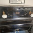 Černé piano s lesklým povrchem a dvěma bílými lampami po stranách. V horní části je oválný dekor s ornamentem uprostřed. Dole na korpusu je nápis "E.…