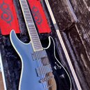 Černá elektrická kytara s dvoma humbuckery, uložená v černém plišovém kufru. Kytara má světlý lem kolem korpusu a hmatník s vkládanými značkami. Vedl…