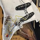 Fotografie zachycuje vnitřní část elektroiky elektrické kytary s bílým pickguardem a hliníkovým stíněním. Jsou vidět tři černé snímače typu single-co…