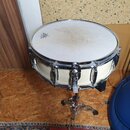 Bílá buben (pravděpodobně snare) na chromovaném stojanu. Na bláně je viditelné logo REMO a značná špína v centru. Korpus je bílý s chromovaným obručo…