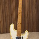 Světlě žlutá elektrická basová kytara s bílým pickguardem a hnedým hmatníkem. Na hlavě je viditelné logo Squier. Nástroj je umístěn na černém stojanu…