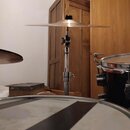 Fotografie zachycuje část bicí sady. V centru je vidět kovový stojan s nasazeným činelem zlatavé barvy. V popředí je vidět horní část bubnu s bílou m…