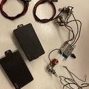 Sada elektroniky pro kytaru obsahující dva černé obdélníkové snímače s nápisem EMG, modrou základnu s rozpojenými součástkami a několik potenciometrů…
