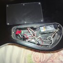 Zadní strana černé kytary s otevřeným přístupem k elektronice. V dutině jsou vidět zapojené součástky, kabely a malý černý modul s červeným konektore…