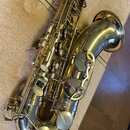 Zlatý saxofon položený na béžovém povrchu. Na těle nástroje jsou viditelné klapy s perleťovými vložkami a kovové mechanismy. Povrch je lesklý s někol…