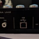 Zadní strana černého audio rozhraní s nápisem U-PHORIA UM2. Jsou zde vidět dva RCA výstupy označené jako OUTPUTS 2 (R) a 1 (L), USB port s odpovídají…