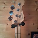Hlava basové kytary z přírodního dřeva s chromovanými ladičkami. Na dřevě je černý nápis „Fender“ a vedle něj svisle text „PRECISION BASS®“ a menší n…