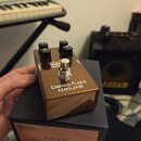Zlatý efekt pedál s nápisy MXR BASSROOM a bass fuzz deluxe. Pedál má čtyři černé ovládací knoflíky a jeden stomečový spínač. Je umístěn na černé krab…