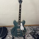 Polodupatá elektrická kytara v modrozelené barvě s bílým pickguardem a chromovaným hardwarem. Na hlavě hmatníku je bílý nápis Epiphone a logo ve tvar…