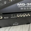 Zadní strana černého efektového pedálu s nápisy NUX MG-30 VERSATILE MODELER. Jsou zde viditelné konektory s popisky INPUT, AUX IN, SEND, RETURN, OUTP…