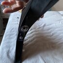 Detailní pohled na hranu černého trupu elektrického nástroje. Je vidět kovová zásuvka pro kabel a malý šroubek v černém povrchu. Nástroj je položen n…