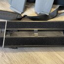 Na fotografii je černý obdélníkový předmět s dvěma širokými pásy černého texturovaného materiálu. V horní části je vidět černý popruh s vyraženým náp…