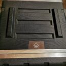 Černý transportní kufr z texturovaného materiálu s kovovými hranami. Na povrchu jsou vyvýšené žebrování a v dolní části je hnědá nálepka s ikonou šip…