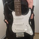 Černá elektrická kytara s bílým pickguardem a hnedým hmatníkem. Nástroj má tři bílé snímače typu single-coil a dva bílé oválné knofíky s nápisy VOLUM…