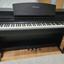Černý digitální piano s klaviaturou a třemi pedály. Na horní části je nápis YAMAHA. Nástroj vykazuje známky běžného používání, jsou vidět drobné oděr…