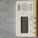 Bílý chromatický ladič značky KORG, model CA 2. Zařízení má digitální displej s hodnotou 440 a tři ovládací tlačítka. Na pravé straně je umístěn žlut…