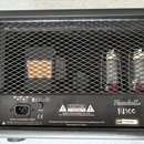 Zadní strana kytarového komba v černém provedení s kovovou mřížkou, za kterou jsou vidět elektronické součástky a čtyři elektrony. Na spodním černém…