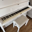 Bílý pianino s klaviatura v krémovém odstínu. Na čelní straně nad klávesami je černý nápis PETROF. K nástroji patří bílá stolička s béžovým polstrová…