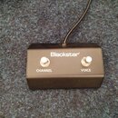 Černý přepínací pedál s logem Blackstar. Na horní straně jsou dvě stříbrná tlačítka s nápisy CHANNEL a VOICE. Z horní části zařízení vychází černý ka…