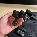 Fotografie zobrazuje čtyři malé černé mikrofony s pěnovými windscreeny, z nichž jeden drží ruka. Na těle mikrofonu je nápis „12 VOLT SOURCE POWER“ a…