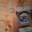 Detail hlavy kytary z přírodního dřeva. Na povrchu je nalepený kulatý stříbrný štítek s nápisy „Fender“ a „60“. Jsou vidět dva chromované ladicí kolí…