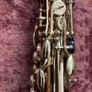 Detailní pohled na zlatý kovový saxofon s mechanismem klapek. Nástroj je v lesklém povrchu bez viditelných značek, log nebo modelových označení. Na p…