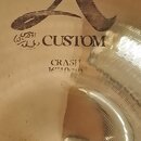 Detail zlatavého kovového činely s viditelným opracováním povrchu. Na ploše je vyryto velké stylizované písmeno A, pod ním nápis CUSTOM a slovo CRASH…
