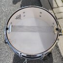 Fotografie znázorňuje snare drum s bílým membránou a chromovaným obručováním. Na membráně je viditelné logo REMO s nápisem Ambassador Coated. Přes ko…