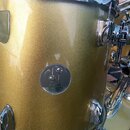 Detail zlatého bubnu s metalickým povrchem a chromovaným kováním. Uprostřed korpusu je oválný kovový štítek s logem dvou kuliček a nápisem SONOR. Pov…