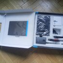 Otevřená modro-bílá krabice s polystyrenovou vložkou, ve které je umístěno příslušenství. Obsahuje šedý přístroj s nápisem SENNHEISER, černý kabel a…