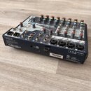 Černý mixovací pult s bílým horním panelem a barevnými ovratovými knoflíky. Na přístroji jsou viditelné nápisy Soundcraft a NOTEPAD-12FX. Na zadní st…