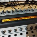 Detailní pohled na ovládací panel elektronického zařízení s displejem. Na oranžovém displeji je text: Setup Menu, LCD Contrast, Factory Def., Jog Whe…