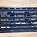 Černý kovový pedalboard s obdélníkovým tvarem a vodorovnými otvory. Povrch je částečně polepený černými sametovými nebo pěnovými pásky. Na nástroji n…