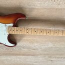 Elektrická kytara s trupem v barevném přechodu od žluté k tmavě hnědé a bílým pickguardem. Na hlavě hmatníku je čitelné logo Fender STRATOCASTER. Nás…