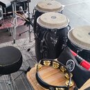 Sestava perkusních nástrojů zahrnující tři kongy s černým dřevěným povrchem a světlými koženými membránami. V popředí je dřevěný cajón, na kterém lež…