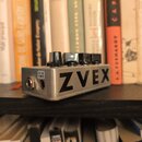 Stříbrný efekt pedál s nápisem ZVEX na boční straně. Na horní části jsou tři černé ovládací knoflíky. V levé části je vidět vstupní konektor a nápis…