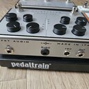 Stříbrný kytarový efekt s černými ovládacími knoflíky, umístěný na černé desce s nápisem pedaltrain. Na zadní straně efektu jsou viditelné nápisy FKT…