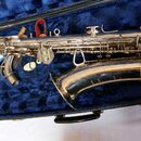 Stříbrný saxofon v otevřeném černém kufru s modrým polstrováním. Nástroj je vybaven kovovými klapy a mechanismy, v kufru jsou vedle něj vidět munční…