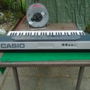 Šedý elektronický klavír s bílými a černými klávesami, na jehož levé straně je nápis CASIO. Nástroj leží na hnědém proutěném stole s zeleným podklade…