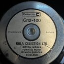 Fotografie zachycuje zadní stranu reproduktoru s kovovým krytem. Na středovém štítku je logo Celestion a označení G12-100. Štítek obsahuje nápisy Imp…