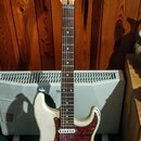 Bílá elektrická kytara typu Stratocaster s hnědým hmatníkem a červeně vzorovaným pickguardem. Na hlavě je viditelné logo Fender. Kytara vykazuje znám…