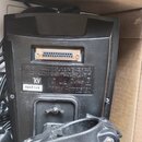 Černý plastový elektronický přístroj v kartonové krabici. Na spodní straně je viditelný vícepinový konektor a štítek s nápisy KV a R6H8149. Povrch pl…