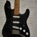 Černá elektrická kytara s tvarem Stratocaster, bílým pickguardem a hnědým javorovým hmatníkem. Nástroj má tři bílé snímače typu single-coil, tři ovlá…