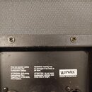 Detail zadní strany reproduktorového boxu s černým povrchem a texturovanou přední mřížkou. Na černém plastovém panelu je bílý nápis Warwick AMPLIFICA…