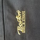 Detailní záběr na černý textilní povrch s zipem. Na látce je žlutým vyšívaným písmem nápis „Boston“ a vedle něj svisle text „BAGS & STRAPS“. Povrch m…