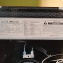 Fotografie detailně zachycuje bílý štítek s technickými specifikacemi na černém povrchu kytarového zesilovače. Štítek obsahuje nápisy GUITAR AMPLIFIE…