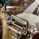 Detailní pohled na snare drum s chromovaným obručem a připevněným pleteným popruhem v bílo-šedé barvě. Na kovovém prvku popruhu je vyražen nápis MADE…