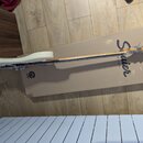 Bílá elektrická kytara položená na hnědé papírové krabici. Na krabici je velký nápis Squier by FENDER a malé logo s textem SQUIER. Nástroj má světlý…